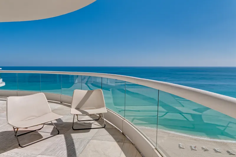 Curved glass balcony with two lounge chairs above a turquoise ocean and beach
