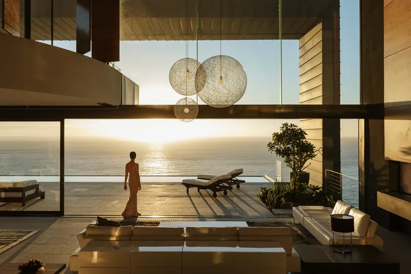 Person standing at a glass wall overlooking the ocean at sunset in a modern living space